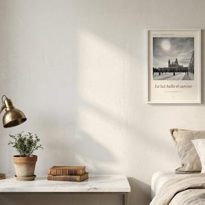 Op de afbeelding: Een ingelijste zwart-witprint met een gebouw en de tekst La luz halla el camino, weergegeven aan een muur naast verschillende kleinere vintage prints, een gedroogd boeket bloemen, een polshorloge en een klein glazen flesje.