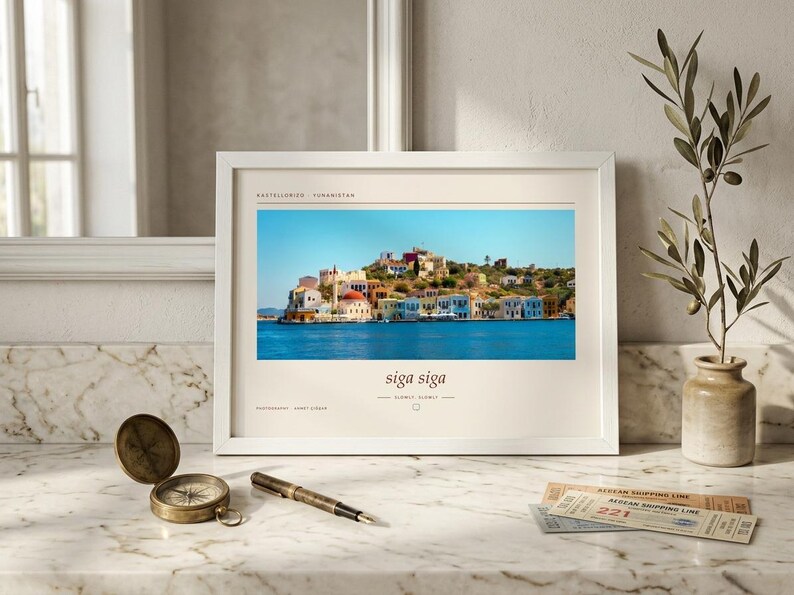 May include: A white framed print featuring a landscape photograph of the Greek island Kastellorizo with colorful buildings by the blue sea. The text reads Kastellorizo, Yunanistan, siga siga, slowly, slowly, and photography by Janet Stiber.