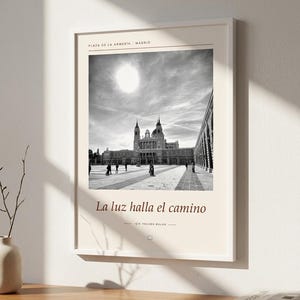 Op de afbeelding: Een ingelijste wandkunstprint met een zwart-witfoto van de Plaza de la Armeria in Madrid. De tekst luidt Plaza de la Armeria - Madrid, La luz halla el camino en IŞIK YOLUNU BULUR, met een klein vierkant icoontje eronder.