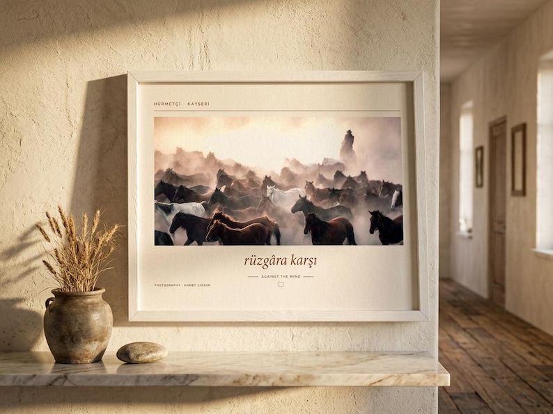 Op de afbeelding: Een ingelijste horizontale wandkunstprint met een foto van een kudde paarden die door stof rent. De tekst luidt HERMET&Ccedil;İ | KAYSERİ, r&uuml;zg&acirc;ra karşı, AGAINST THE WIND en PHOTOGRAPHY AHMET ZİREK. De lijst is gemaakt van lichtgekleurd hout.