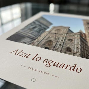 Op de afbeelding: Een rechthoekige print op gebroken wit papier met een foto van de gevel van de kathedraal van Florence. De tekst "Alza lo sguardo" is in bruin gedrukt, gevolgd door "BAŞINI KALDIR" en een klein vierkant icoontje eronder.