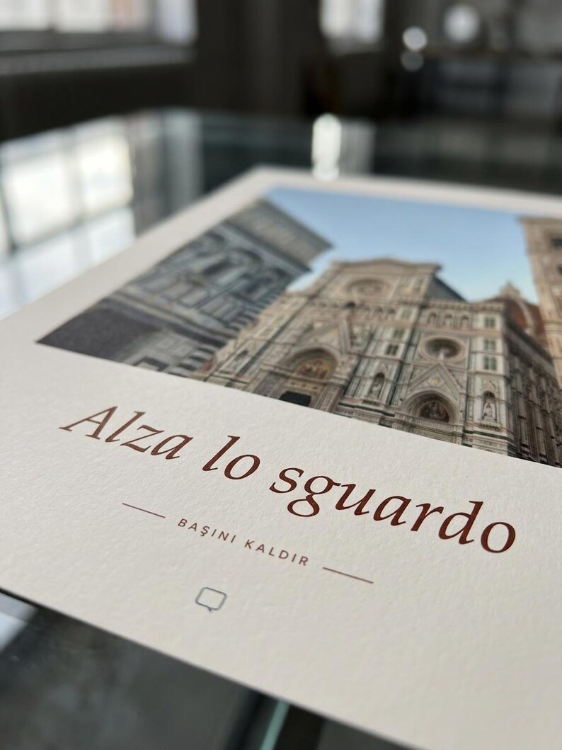 Op de afbeelding: Een rechthoekige print op gebroken wit papier met een foto van de gevel van de kathedraal van Florence. De tekst "Alza lo sguardo" is in bruin gedrukt, gevolgd door "BAŞINI KALDIR" en een klein vierkant icoontje eronder.