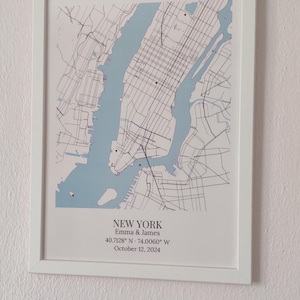 May include: A framed map print of New York City featuring light blue water and black street lines. Below the map, the text reads: NEW YORK, Emma & James, 40.7128&deg; N &middot; 74.0060&deg; W, October 12, 2024. The print is housed in a simple white frame.