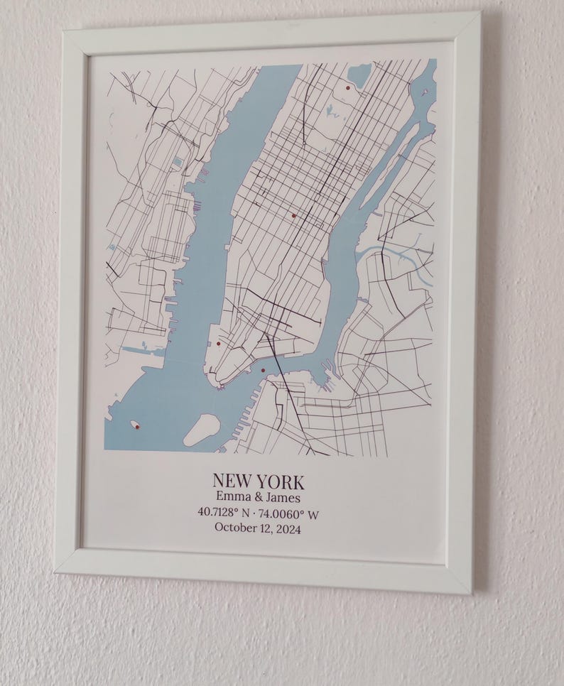 May include: A framed map print of New York City featuring light blue water and black street lines. Below the map, the text reads: NEW YORK, Emma & James, 40.7128&deg; N &middot; 74.0060&deg; W, October 12, 2024. The print is housed in a simple white frame.