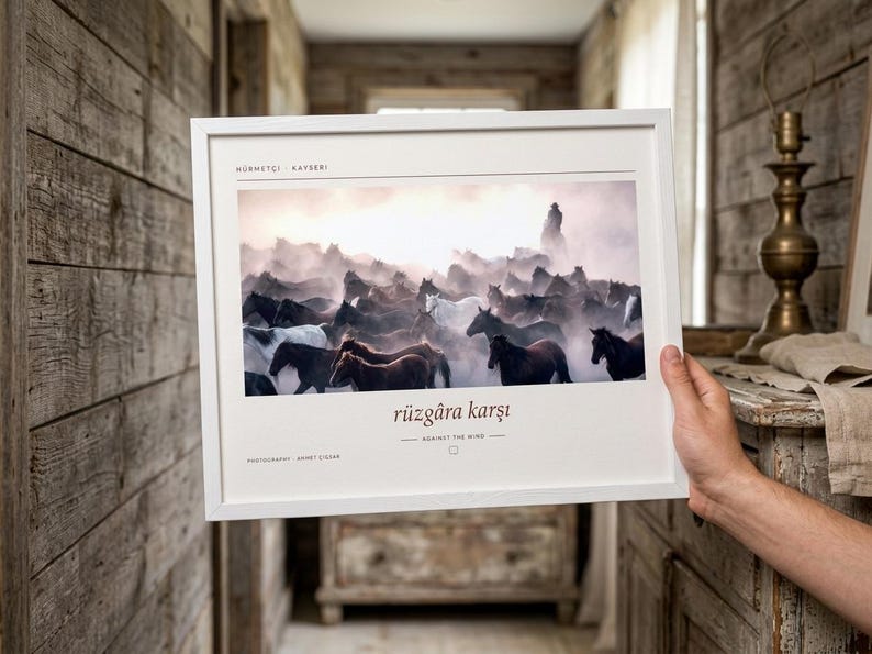 Op de afbeelding: Een wit ingelijste kunstdruk met een foto van een kudde paarden die bij zonsondergang door stof rent. De tekst H&Uuml;RMET&Ccedil;İ KAYSERİ staat bovenaan en r&uuml;zg&acirc;ra karşı AGAINST THE WIND staat onder de afbeelding gedrukt.