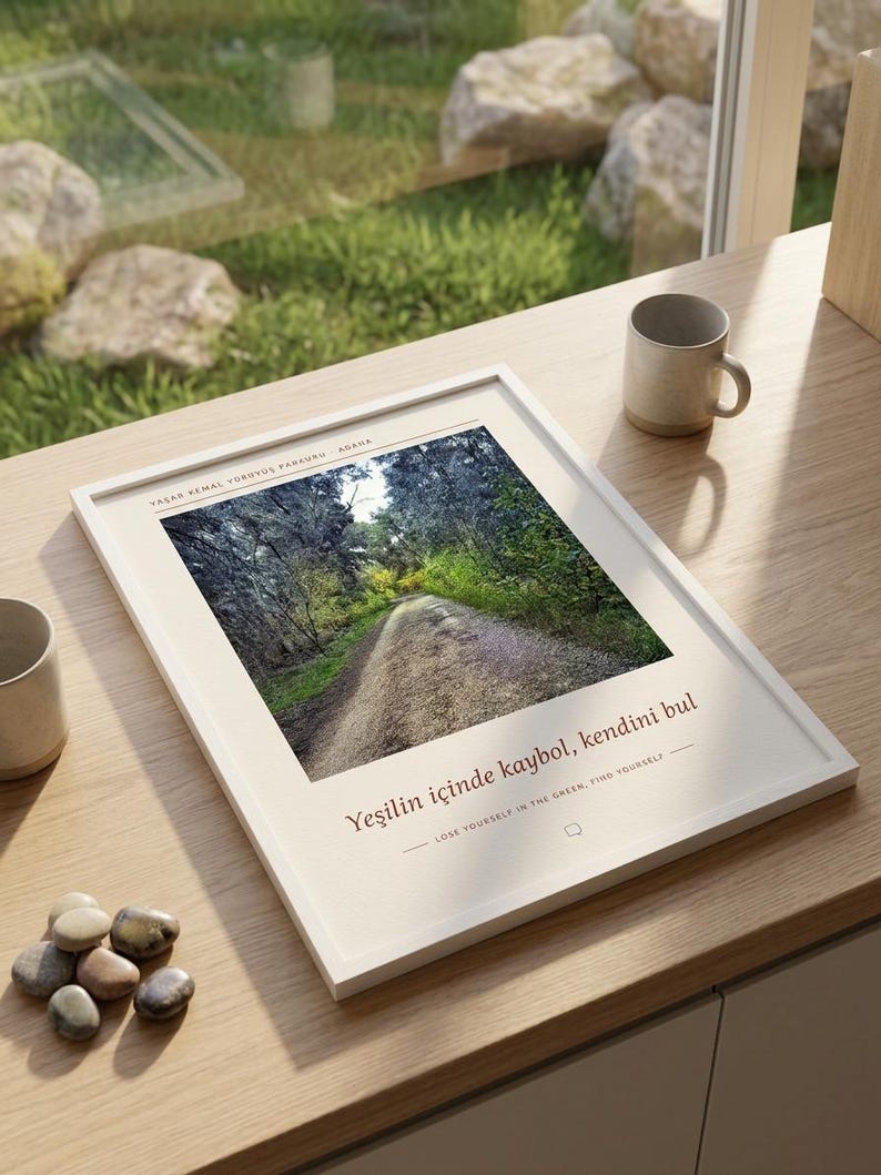 May include: A white framed wall art print featuring a photograph of a forest path. The text reads: YAŞAR KEMAL Y&Uuml;R&Uuml;Y&Uuml;Ş PARKURU ADANA, Yeşilin i&ccedil;inde kaybol, kendini bul, and LOSE YOURSELF IN THE GREEN, FIND YOURSELF. The print is displayed on a wooden surface.