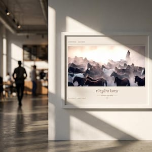 Op de afbeelding: Een ingelijste horizontale wandkunstprint met een foto van een kudde paarden die bij zonsondergang door stof rent. De tekst "H&uuml;rmet&ccedil;i - Kayseri" staat bovenaan en "r&uuml;zg&acirc;ra karşı" met "AGAINST THE WIND" staat onder de afbeelding gedrukt.