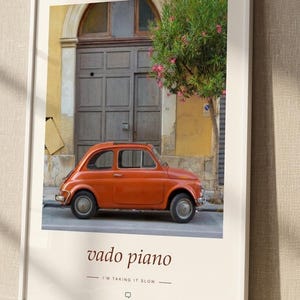 May include: A framed wall art print featuring a vintage orange car parked in front of a grey door and a flowering tree. The text reads SICILIA ITALIA at the top and vado piano I'M TAKING IT SLOW at the bottom, set against a cream background.