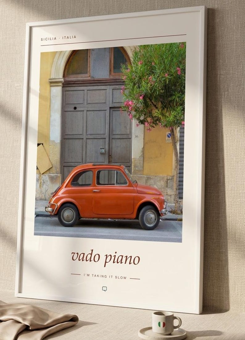 May include: A framed wall art print featuring a vintage orange car parked in front of a grey door and a flowering tree. The text reads SICILIA ITALIA at the top and vado piano I'M TAKING IT SLOW at the bottom, set against a cream background.