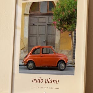 May include: A framed wall art print featuring a vintage orange car parked in front of a wooden door and a flowering tree in Sicily, Italy. The text at the top reads SICILIA ITALIA and the bottom text reads vado piano I'M TAKING IT SLOW.