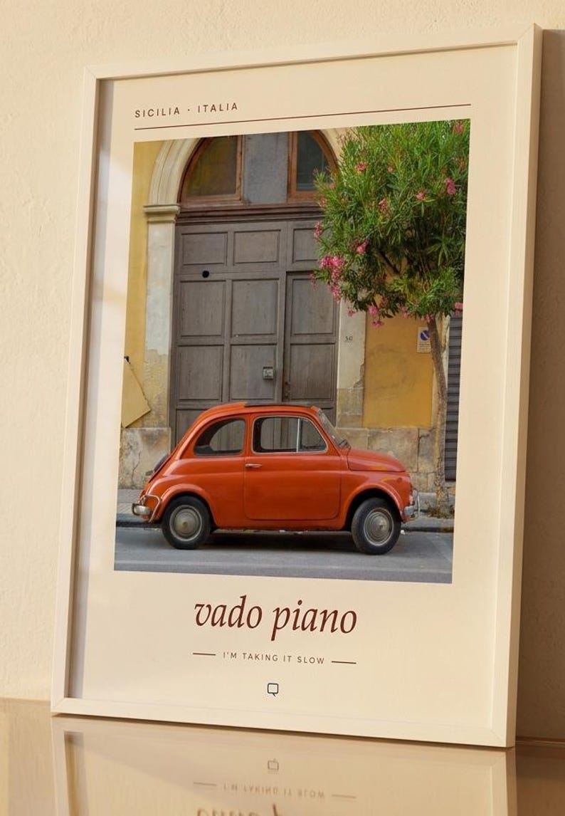 May include: A framed wall art print featuring a vintage orange car parked in front of a wooden door and a flowering tree in Sicily, Italy. The text at the top reads SICILIA ITALIA and the bottom text reads vado piano I'M TAKING IT SLOW.