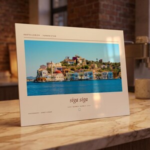 May include: A rectangular white print featuring a landscape photograph of the Greek island Kastellorizo with colorful buildings by the blue sea. The text reads KASTELLORIZO - YUNANISTAN, siga siga, SLOWLY, SLOWLY, and PHOTOGRAPHY - AHMET COŞAR.