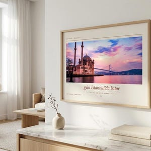May include: A rectangular wall art print in a light wood frame featuring a photograph of the Ortak&ouml;y Mosque in Istanbul at sunset. The print includes the text "g&uuml;n İstanbul'da batar" and "THE SUN SETS IN ISTANBUL" in a clean, modern font.