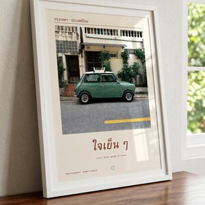 Pode incluir: Uma impress&atilde;o de arte emoldurada com uma fotografia de um carro compacto vintage verde-s&aacute;lvia estacionado numa rua em frente a um edif&iacute;cio. A impress&atilde;o inclui o texto tailand&ecirc;s "กรุงเทพฯ &middot; ประเทศไทย" no topo e "ใจเย็น ๆ" com a tradu&ccedil;&atilde;o inglesa "EASY DOES IT" abaixo.
