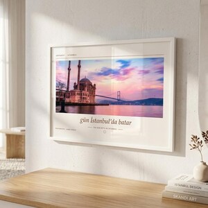 May include: A rectangular wall art print in a white frame featuring a photograph of the Ortak&ouml;y Mosque in Istanbul at sunset. The image displays a pink and blue sky over the Bosphorus bridge and water, with the text g&uuml;n İstanbul'da batar below.