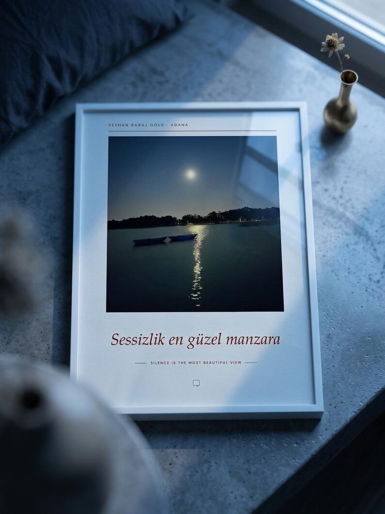 Puede incluir: Una l&aacute;mina de arte enmarcada en blanco con una fotograf&iacute;a de paisaje nocturno de un lago con dos barcas y una luna llena. El texto dice SEYHAN BARAJ GOLU ADANA, Sessizlik en guzel manzara y SILENCE IS THE MOST BEAUTIFUL VIEW con un icono.