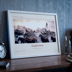 May include: A rectangular white-framed wall art print featuring a photograph of a herd of horses running through dust. The print includes the text "H&Uuml;RMET&Ccedil;İ KAYSERİ" at the top and "r&uuml;zg&acirc;ra karşı" with "AGAINST THE WIND" and "PHOTOGRAPHED AHMET Zİ&Ccedil;AR" below.
