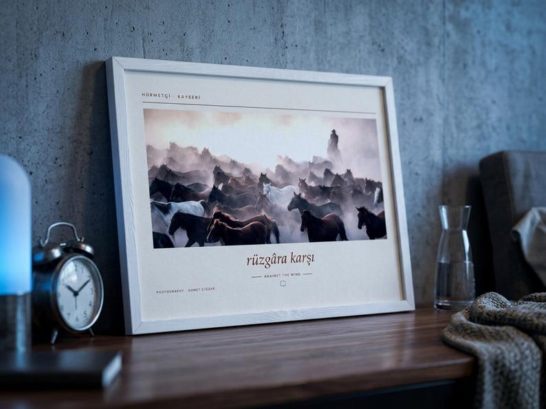 May include: A rectangular white-framed wall art print featuring a photograph of a herd of horses running through dust. The print includes the text "H&Uuml;RMET&Ccedil;İ KAYSERİ" at the top and "r&uuml;zg&acirc;ra karşı" with "AGAINST THE WIND" and "PHOTOGRAPHED AHMET Zİ&Ccedil;AR" below.