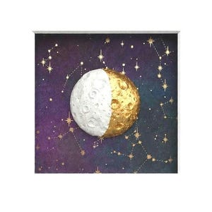 Puede incluir: Una pieza de arte mural cuadrada con una luna tridimensional texturizada centrada sobre un fondo azul y morado oscuro. La luna está dividida en un lado blanco con cráteres y un lado dorado metálico, rodeada de constelaciones de pan de oro.
