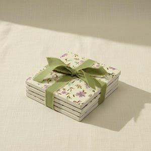 Puede incluir: Una pila de cuatro posavasos cuadrados con fondo crema y un patrón repetitivo de pequeñas flores moradas y verde azulado con tallos marrones. Los posavasos están atados con una cinta de color verde salvia con bordes deshilachados.