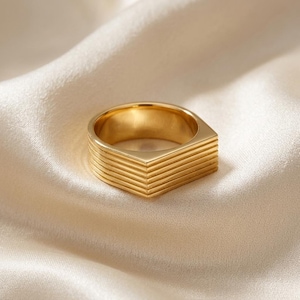 Puede incluir: Un anillo de tono dorado que presenta una punta prominente en forma de chevrón y una serie de estrías horizontales y paralelas que envuelven la banda. La superficie metálica tiene un acabado pulido, creando un aspecto reflectante y geométrico.
