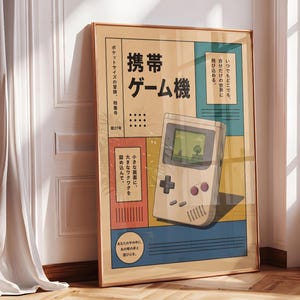Puede incluir: Un póster enmarcado que presenta la ilustración de una consola de juegos portátil de color beige con pantalla verde, una cruceta negra y dos botones rojos. El diseño incluye texto en japonés y bloques de color azul, rojo y amarillo.