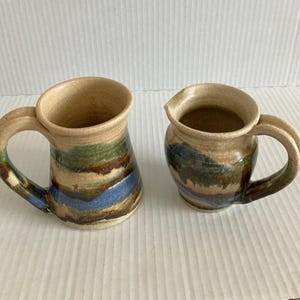 May include: A ceramic mug and a matching creamer pitcher, both featuring a tan base color with horizontal bands of green, brown, and blue glaze. The mug has a tall, slightly flared shape with a side handle, while the pitcher has a rounded body.