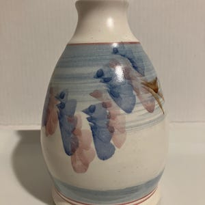 May include: A ceramic vase with a speckled off-white base, featuring horizontal light blue bands and a hand-painted floral motif in shades of blue and muted pink. The vase has a narrow neck, a rounded body, and a flared base with thin pink lines.