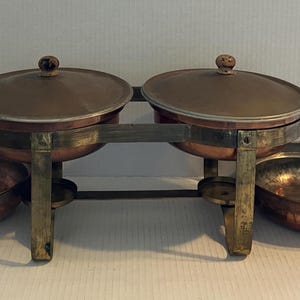 May include: A copper chafing dish set featuring two round lidded pots supported by a brass-toned metal stand, flanked by two matching copper skillets with wooden handles. The metallic surfaces show a warm, aged patina throughout the entire assembly.