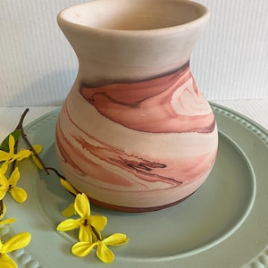 May include: A rounded ceramic vase with a matte finish, featuring a swirling marbled pattern of muted pink, terracotta, and cream tones. The vessel has a wide, flared opening and a smooth, curved body that tapers toward the base.