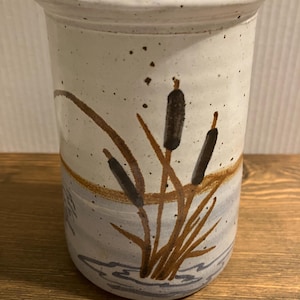 May include: A cylindrical ceramic vase with a speckled off-white upper section and a light blue lower section. The exterior features a hand-painted design of brown cattails and green reeds emerging from stylized blue water ripples at the base.