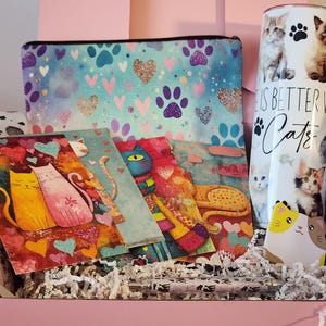May include: A gift box containing a zippered pouch with a blue and purple paw print pattern, a tall tumbler with cat photos and the text "Life is better with cats", two illustrated cat greeting cards, a cat-shaped sticky note, and a patterned pen.