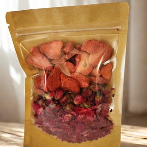 May include: A stand-up pouch with a clear front window and a brown paper back, filled with a mixture of dried red strawberry slices, small dried rose buds, and loose dried rose petals layered within the transparent section of the packaging.
