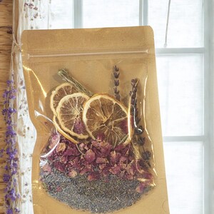 May include: A brown kraft paper pouch with a clear plastic window containing a mixture of dried botanical ingredients, including three circular dried orange slices, purple lavender sprigs, and a base layer of dried pink rose petals and lavender.