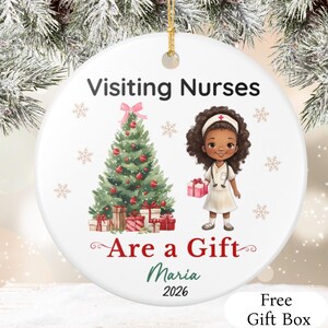 May include: A round white ceramic ornament featuring a Christmas tree with red ornaments and gifts, a nurse illustration with a stethoscope and gift box, and the text Visiting Nurses Are a Gift, Maria, 2026. It includes a gold hanging string.