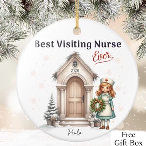 Pode incluir: Um ornamento redondo de cerâmica branca com o texto Best Visiting Nurse Ever, uma ilustração de uma casa com o ano 2026, uma enfermeira com uma guirlanda e o nome Paula. O ornamento inclui um cordão dourado para pendurar no topo.