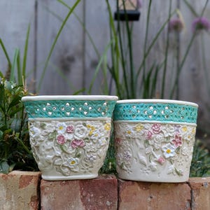 May include: Two off-white ceramic planters featuring a raised floral relief pattern with pink, yellow, and white flowers. Each planter has a wide, turquoise-colored lattice-style rim with a repeating diamond cutout design around the top edge.