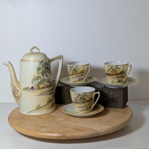 May include: A porcelain tea set featuring a tall teapot and three matching teacups with saucers. The cream-colored ceramic pieces are decorated with a hand-painted landscape scene depicting green trees, grassy fields, and small purple accents.