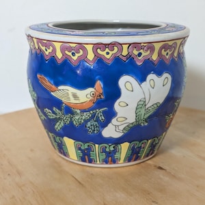 May include: A round ceramic planter featuring a vibrant blue background decorated with a colorful bird perched on a branch and a large white butterfly. The rim and base are adorned with yellow and pink geometric patterns and intricate borders.