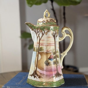 May include: A porcelain coffee pot featuring a hand-painted landscape scene with tall trees, a small house, and a sunset sky. The pot has a green rim, a gold-trimmed handle, and a matching lid with a decorative finial, all finished with gold accents.