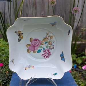 May include: A square white porcelain bowl with scalloped edges and a gold rim. The centre features a floral arrangement with pink roses and green leaves. The interior and sides are decorated with various colourful butterflies and small insects.