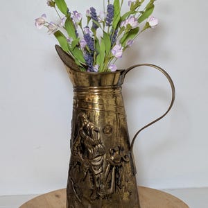 May include: A tall brass pitcher featuring an embossed relief scene of a person working, a curved handle, and a wide spout. The pitcher is filled with artificial lavender and light purple flowers with green stems, creating a decorative arrangement.
