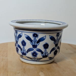 May include: A white ceramic planter featuring a repeating blue floral pattern around the exterior. The bowl-shaped vessel has a wide, flat rim decorated with a geometric border and a slightly flared base, showcasing a classic blue and white design.