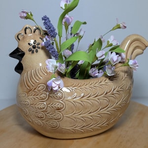 May include: A tan ceramic chicken-shaped planter featuring embossed feather patterns on the body and a dark brown beak. The planter is filled with artificial green stems, small light purple flowers, and textured purple floral spikes.