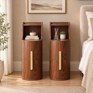 May include: Two cylindrical bedside tables made of dark brown wood with visible grain. Each table features an open top shelf and a lower cabinet with a vertical gold-toned metal handle. The tables are positioned side by side on a light wood floor.