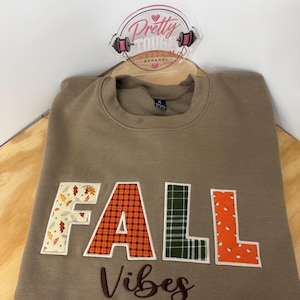 May include: A tan crewneck sweatshirt featuring the word FALL in large, fabric-appliqued letters with various patterns including autumn leaves, orange plaid, green plaid, and pumpkins. The word Vibes is embroidered in dark brown script below.