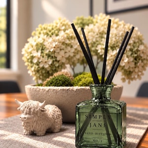 May include: A square, translucent green glass reed diffuser bottle with black reeds. The label reads SIMPLY JANE REED DIFFUSER NO. 1. Beside it sits a small, cream-coloured figurine of a shaggy, horned highland cow.
