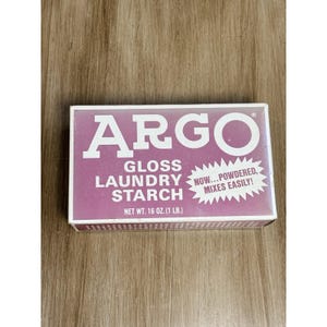 以下が含まれることがあります： 紫色の背景に白い文字が書かれたArgo Gloss Laundry Starchの長方形の箱。前面にはブランド名、製品タイプ、および白い星形のバースト内に「NOW...POWDERED, MIXES EASILY!」というテキストが表示されています。正味重量は453グラムです。