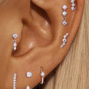 May include: A collection of silver-toned ear piercings featuring clear, round, and marquise-cut stones. The set includes multiple stud earrings, small hoops, and dangling charms arranged along the ear cartilage, lobe, and tragus areas.