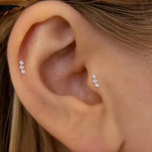 May include: Two silver-toned ear piercings, each featuring a vertical row of three small, clear, round-cut stones. One earring is positioned on the upper cartilage, while the other is placed on the tragus, both set in a metallic base.